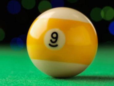 A yellow and white billiard ball with the number nine on it lying on a pool table.