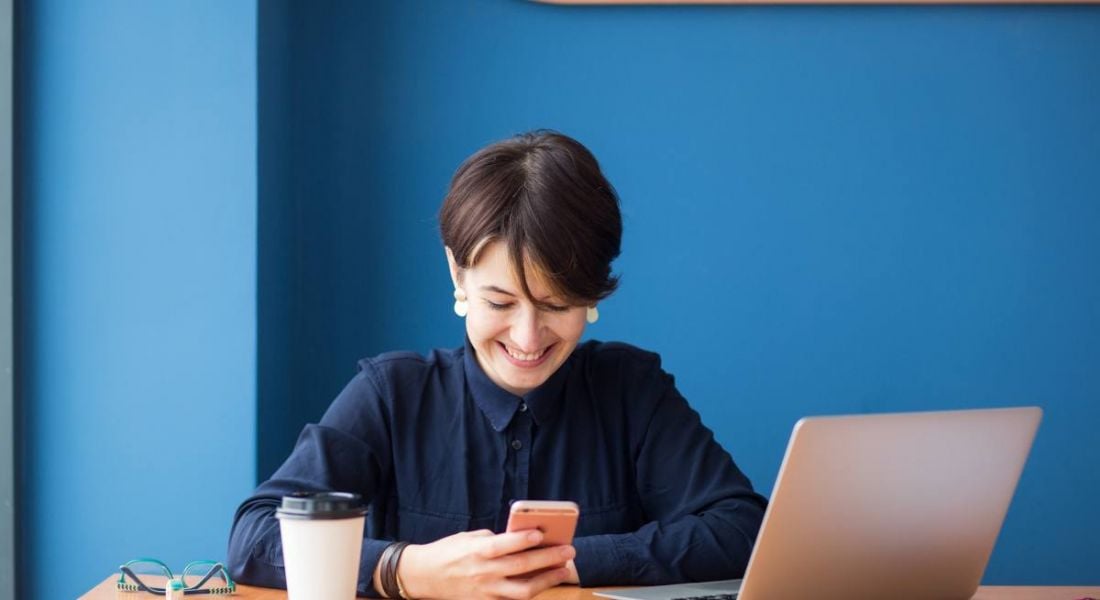 Businesswoman sitting at a table with a laptop, phone and coffee cup beside her. A bright blue wall is behind her.