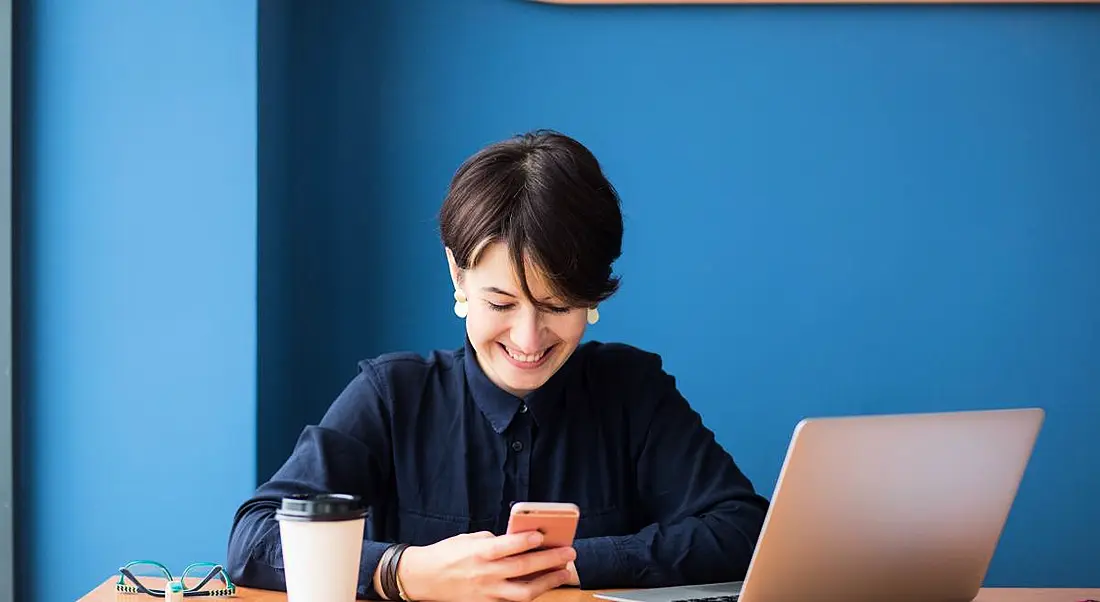 Businesswoman sitting at a table with a laptop, phone and coffee cup beside her. A bright blue wall is behind her.