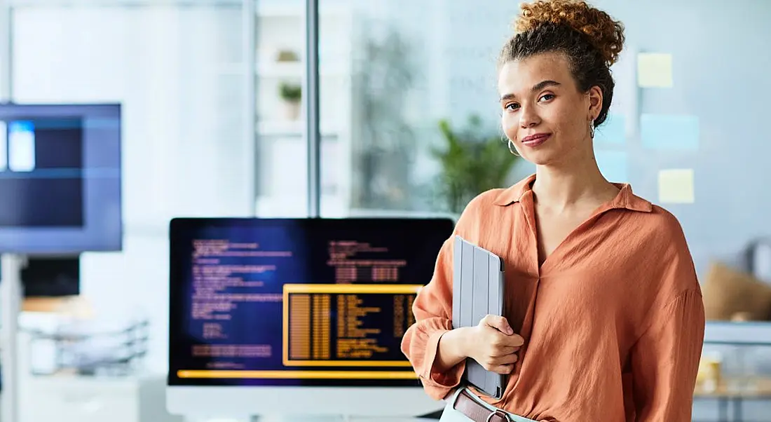 Young woman standing in a modern office in front of a large computer monitor displaying lines of code.