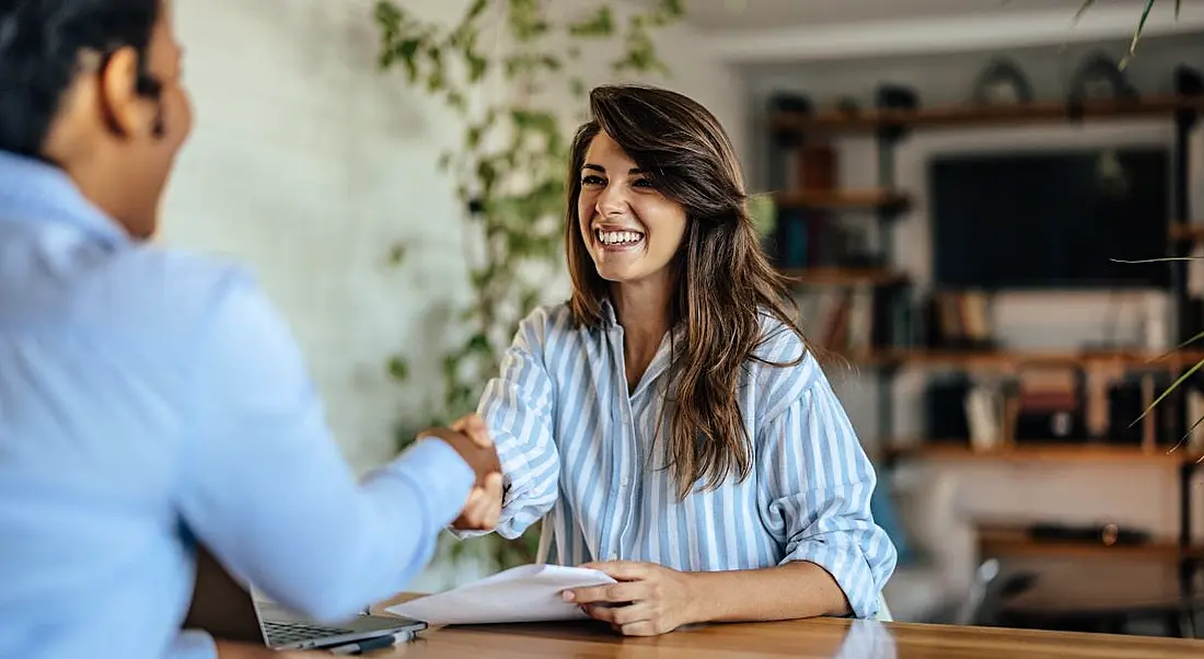 A woman shakes hands with another woman across a table while sitting down to be interviewed.