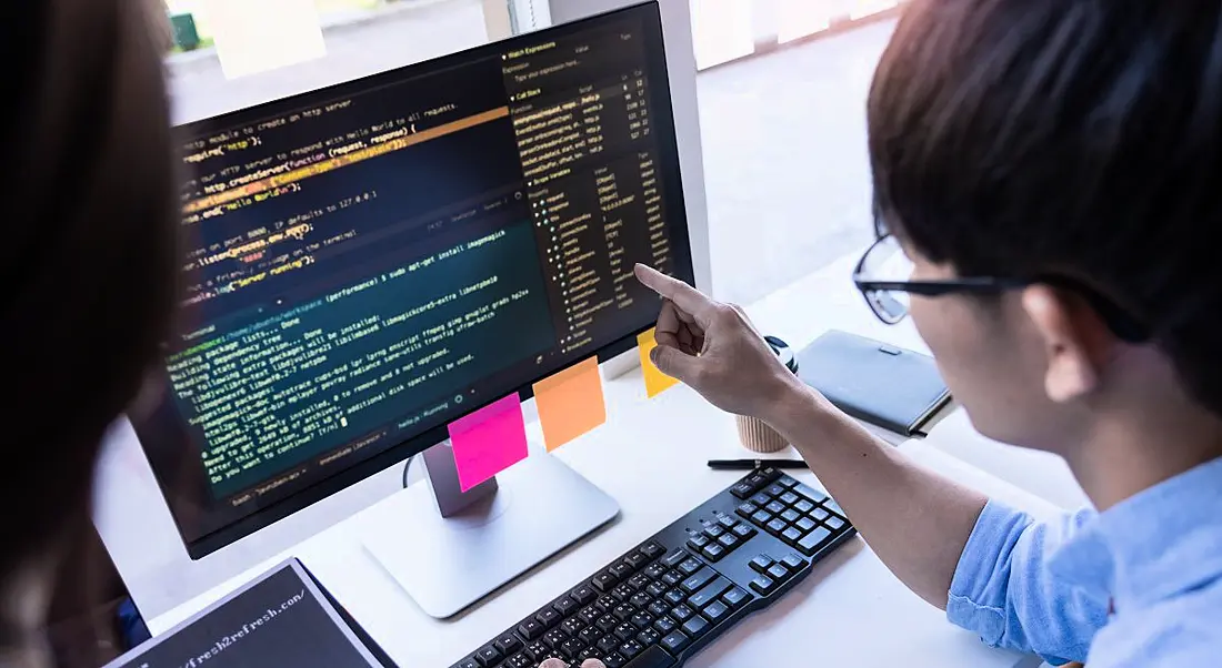 A man sitting at a desk pointing at a screen with a lot of code on it.