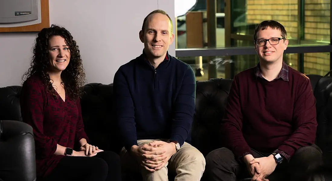 Three people who work at SciLeads sitting on a dark coloured leather sofa in a dimly lit room with a wall and glass behind them.