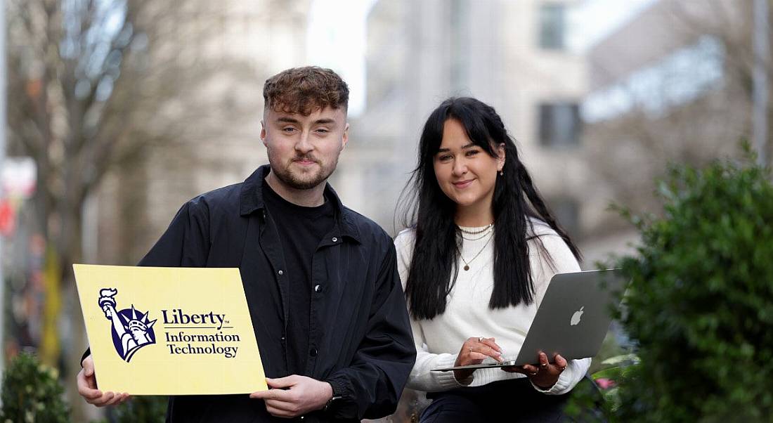 A young man and woman standing outside a set of brown buildings holding a laptop and a sign with Liberty IT on it.