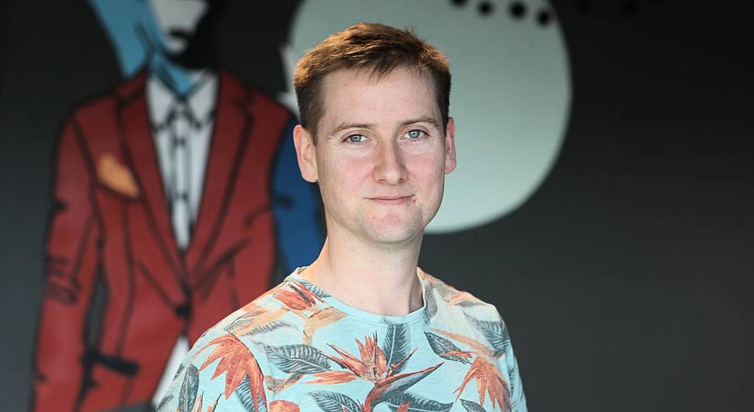 A young man in a colourful t-shirt smiling at the camera in the Zalando office.