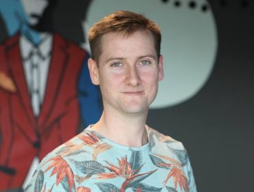 A young man in a colourful t-shirt smiling at the camera in the Zalando office.