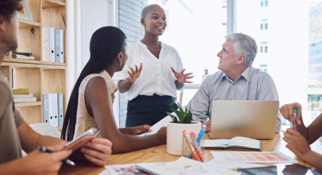 A group of people sitting at a table in a brainstorming session, while listening to one woman who is standing, showing off her soft skills.