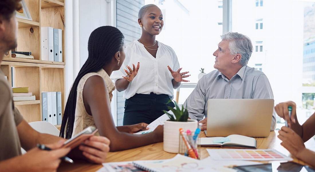 A group of people sitting at a table in a brainstorming session, while listening to one woman who is standing, showing off her soft skills.