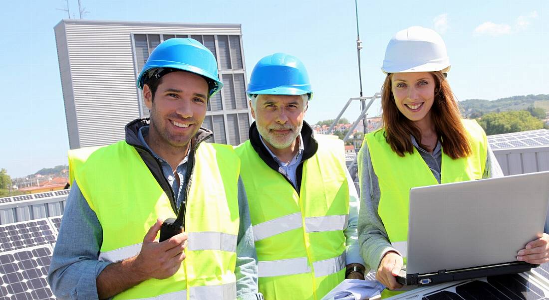 Three engineers in protective gear standing on a rooftop outside. One has a laptop.