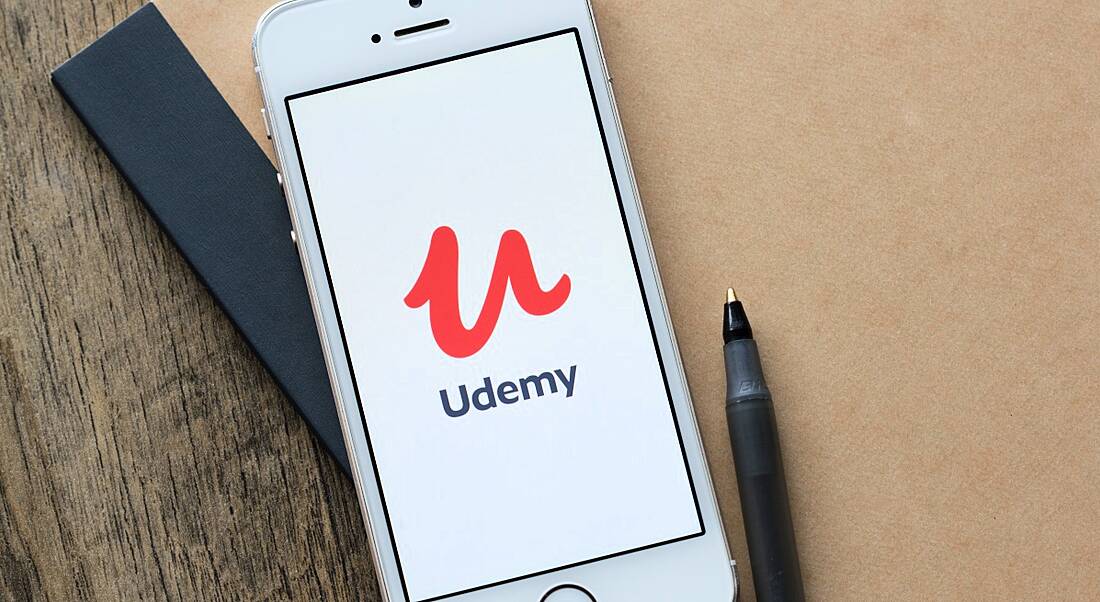 A mobile phone with the Udemy app open on it lying on a notebook with a pen beside it.