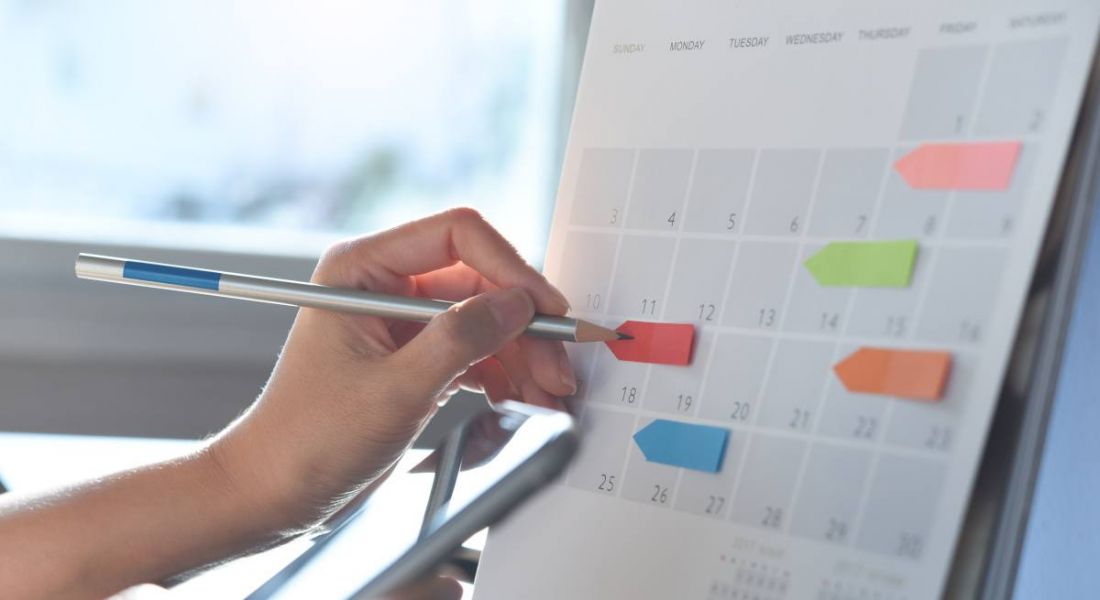 A paper calendar of a month with different coloured notes on certain days. A person's hand is writing on one of these notes.