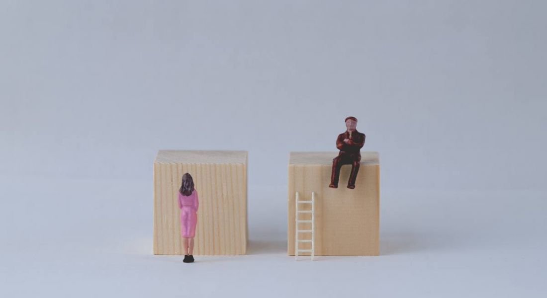 A man figurine sitting on a wooden block with a ladder beside him with a woman figurine standing in front of a wooden block. The cartoon represents the gender pay gap.