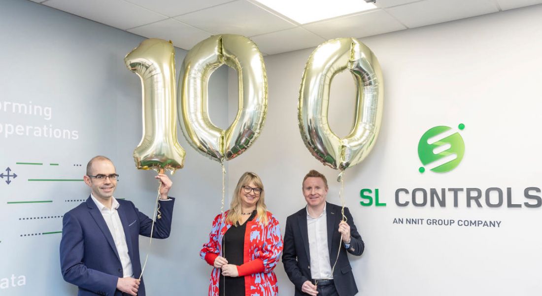 Two men and a woman stand in an office holding gold foil balloons that spell 100. The SL Controls logo is on the wall beside them.