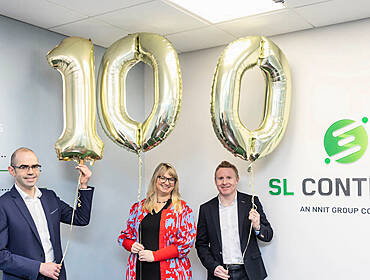Two men and a woman stand in an office holding gold foil balloons that spell 100. The SL Controls logo is on the wall beside them.