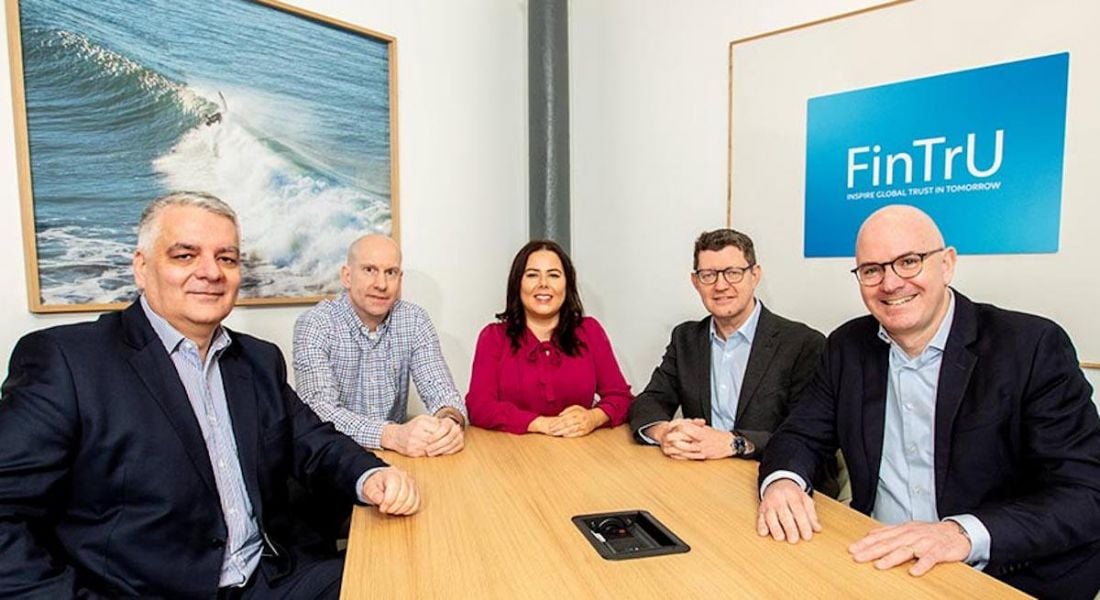 Four men and one woman sit around a boardroom table smiling at the camera. A sign that says FinTru is on the wall behind them.