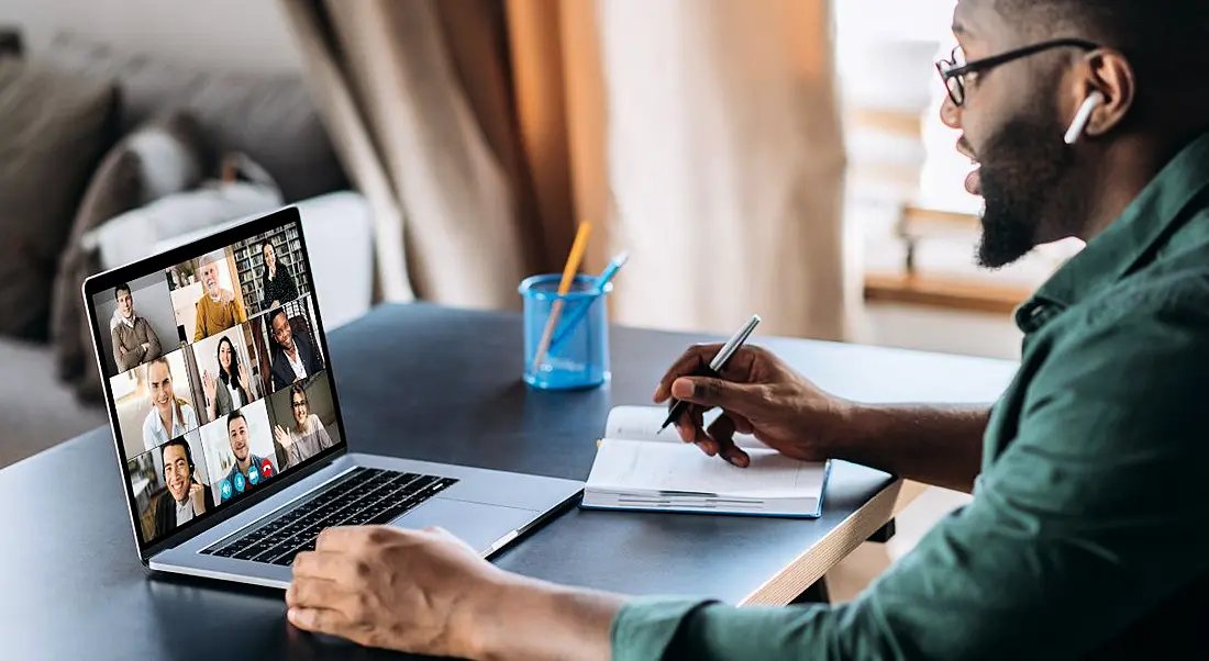 A man on sitting at a desk with a laptop. He is on a video call while taking notes, symbolising remote work.