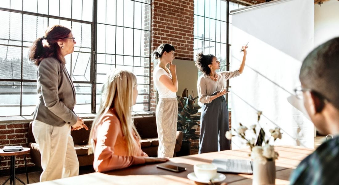 A meeting room filled with five people, all looking at a long white sheet of paper, with one of the people holding a pen near it, contemplating writing on it. The sun shines brightly into the room through large windows.