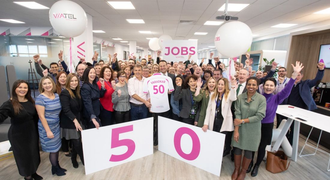 Taoiseach Leo Varadkar stands at the centre of a group of Viatel staff. he is holding a cardboard jersey cutout with his name and the number 50 on the back and there is a big sign with 50 on it in pink writing. They are in Viatel's Blanchardstown office.