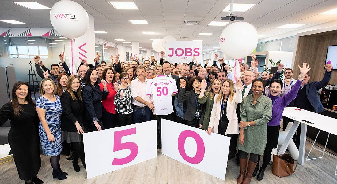 Taoiseach Leo Varadkar stands at the centre of a group of Viatel staff. he is holding a cardboard jersey cutout with his name and the number 50 on the back and there is a big sign with 50 on it in pink writing. They are in Viatel's Blanchardstown office.