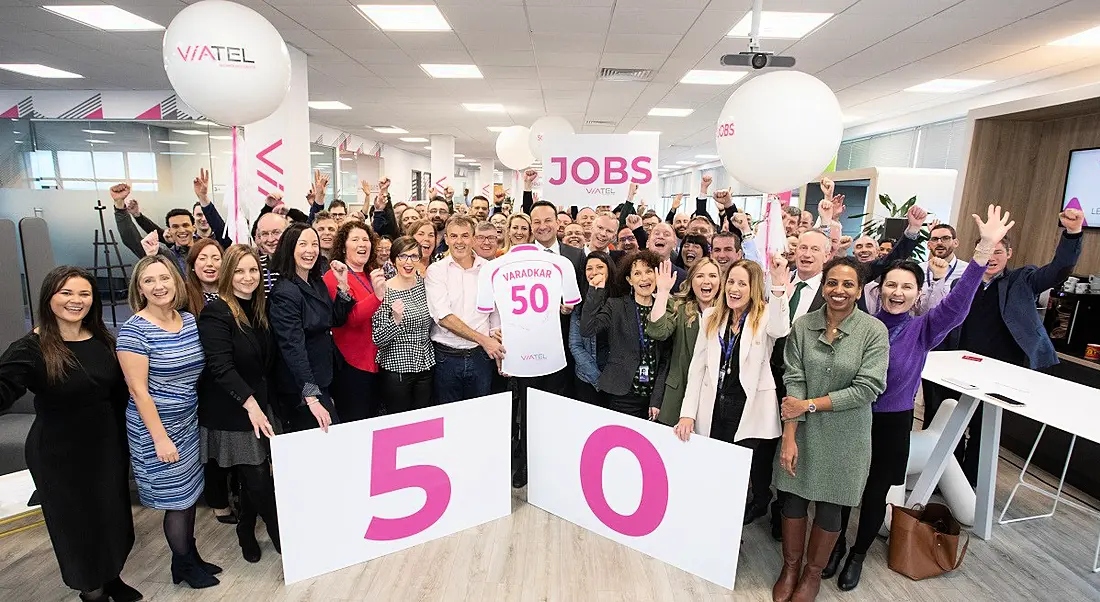 Taoiseach Leo Varadkar stands at the centre of a group of Viatel staff. he is holding a cardboard jersey cutout with his name and the number 50 on the back and there is a big sign with 50 on it in pink writing. They are in Viatel's Blanchardstown office.