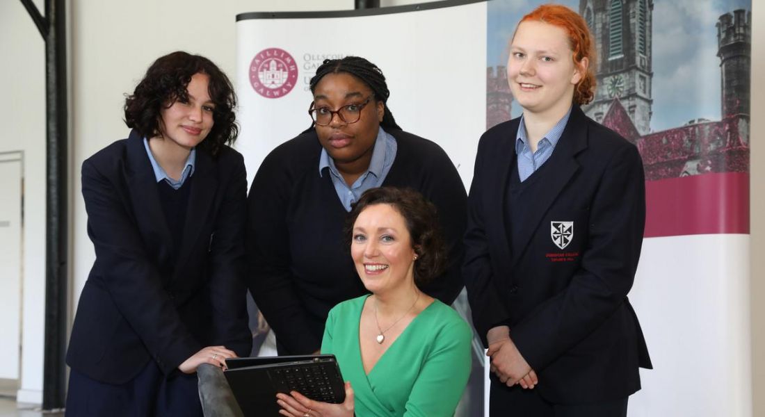 A woman sitting down holding a laptop computer with three teenagers standing behind her in a group.