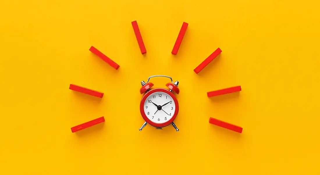 A red alarm clock with red dominoes arranged around it in a semicircle. Yellow background.