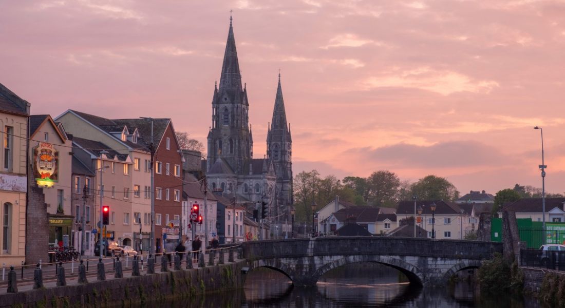 View of St Finbarr's Cathedral across the River Lee at dusk with buildings around it.