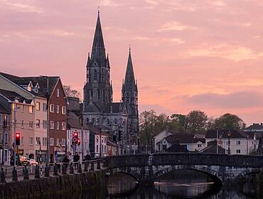 View of St Finbarr's Cathedral across the River Lee at dusk with buildings around it.