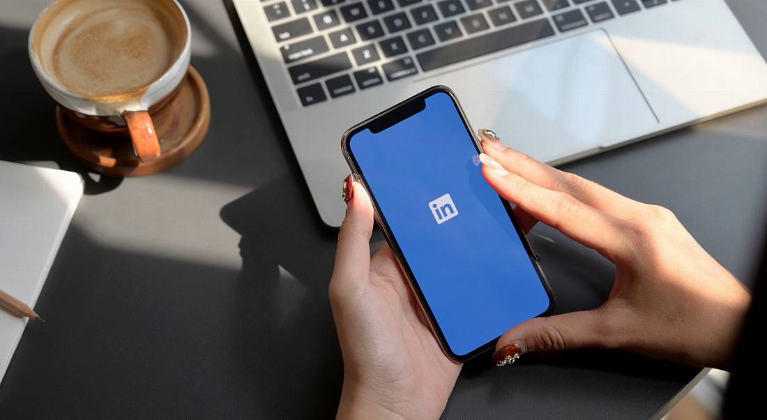 Person's hands holding a phone with LinkedIn branding on it as they use a laptop. There is a coffee beside them.