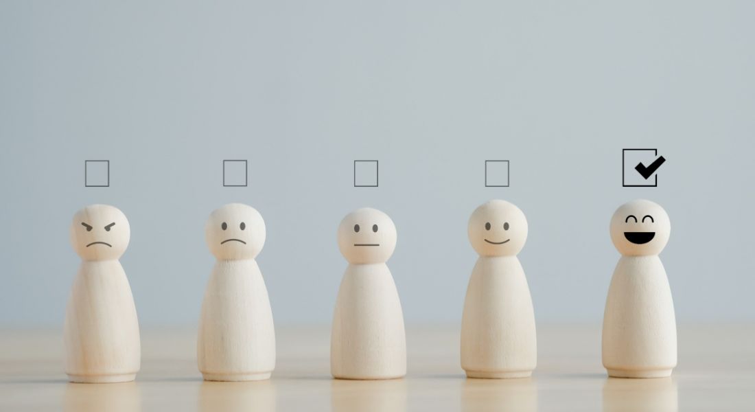 A row of wooden figurines with facial expressions ranging from sad to happy and check boxes over their heads with the box above the happy figure's head checked.