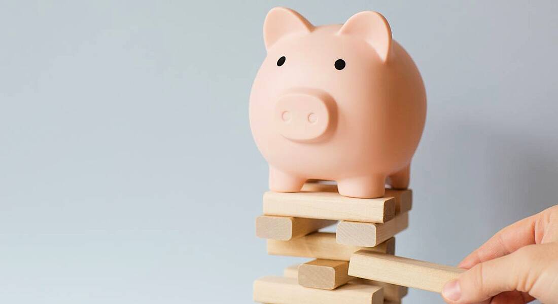 Piggy bank sitting on top of a Jenga tower of bricks with a person's hand pulling a brick from the tower.