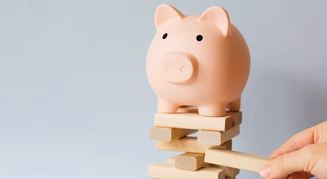 Piggy bank sitting on top of a Jenga tower of bricks with a person's hand pulling a brick from the tower.