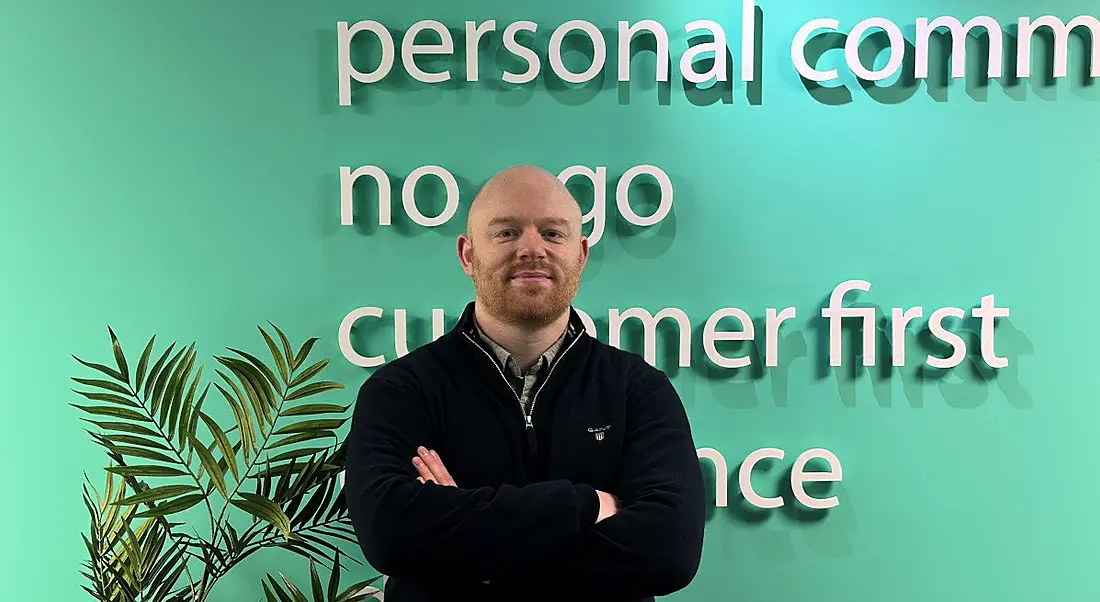 A man stands with his arms folded smiling at the camera with a green wall behind him. He is Dan McElligott, a technical lead at Version 1.