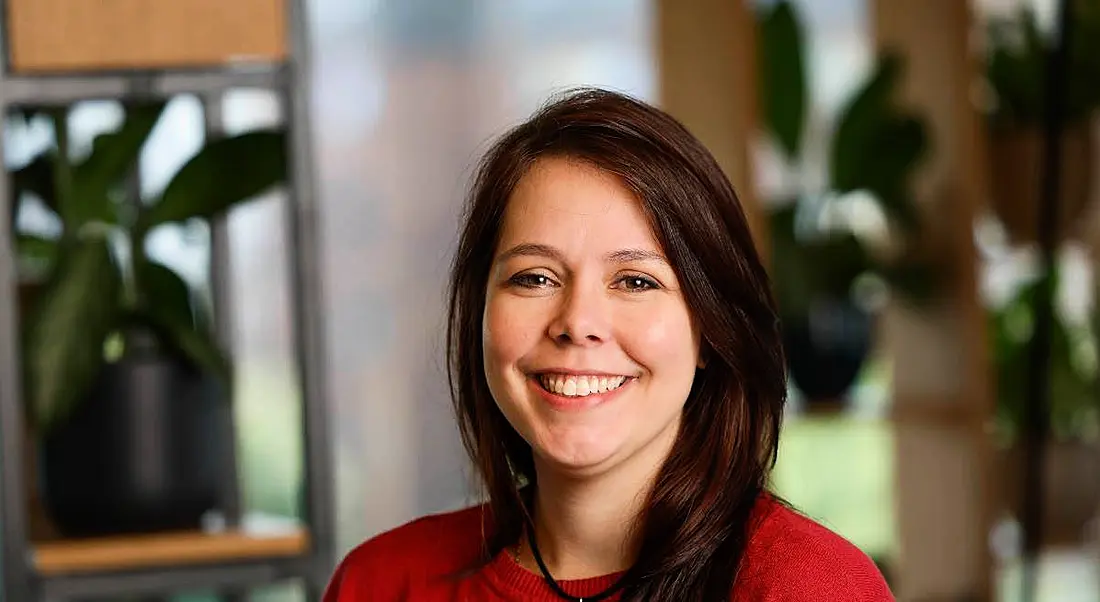 A young woman with dark hair wearing red. She smiles at the camera while standing in a bright office.