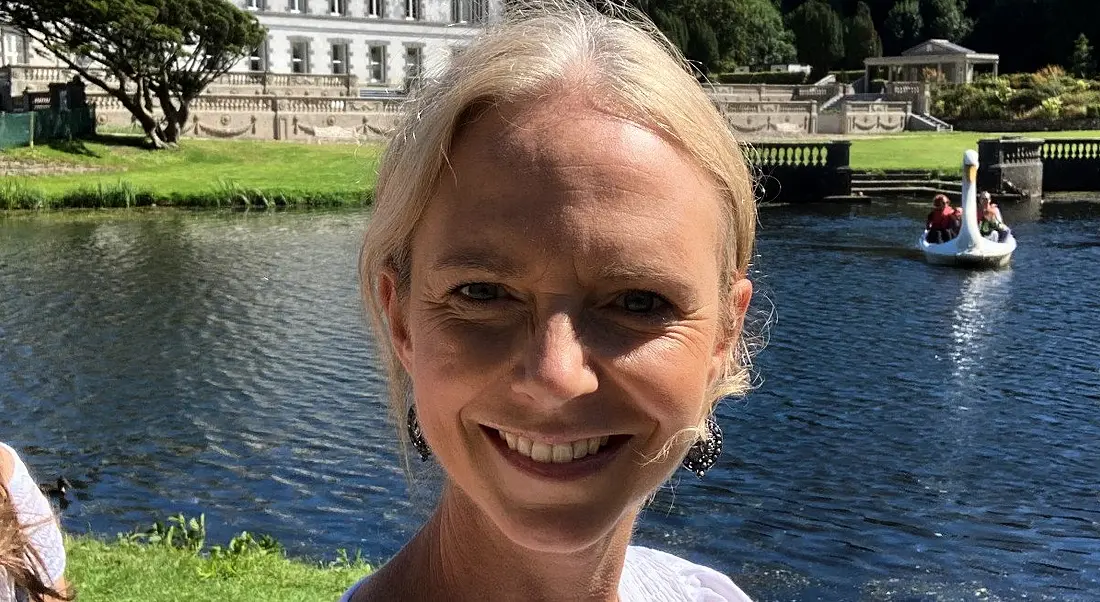 A woman smiles at the camera with a lake in the background. She is Breda O'Callaghan.