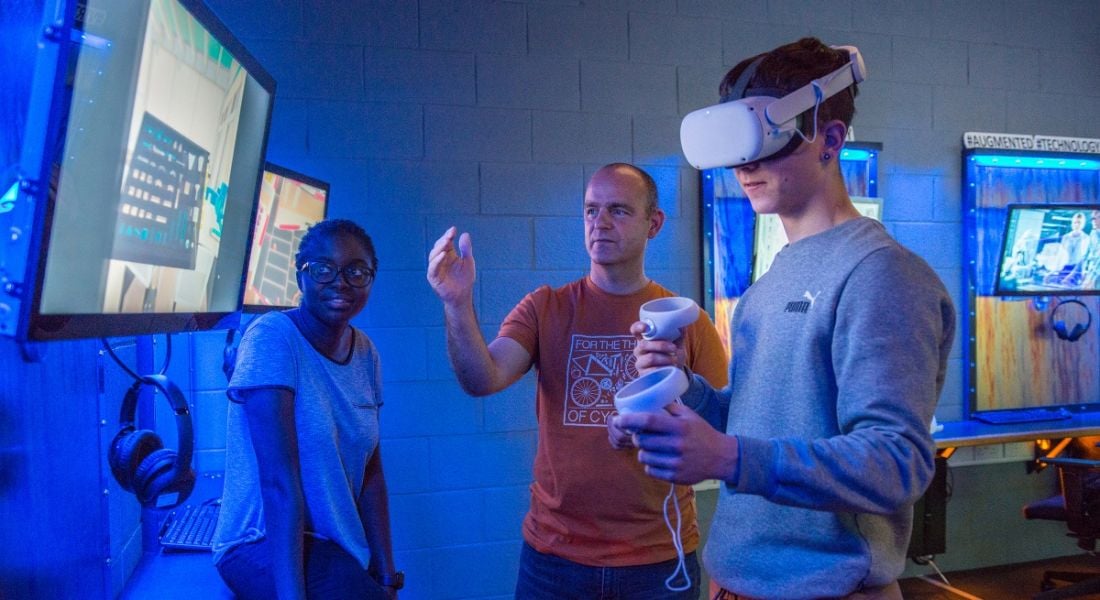 Two engineering students using VR headsets and standing in front of a screen in a classroom. They are accompanied by a lecturer.