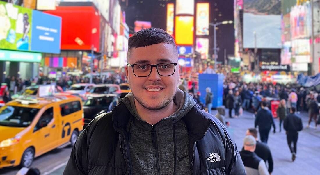 A young man with glasses smiles at the camera. In the background is a very busy city at night.