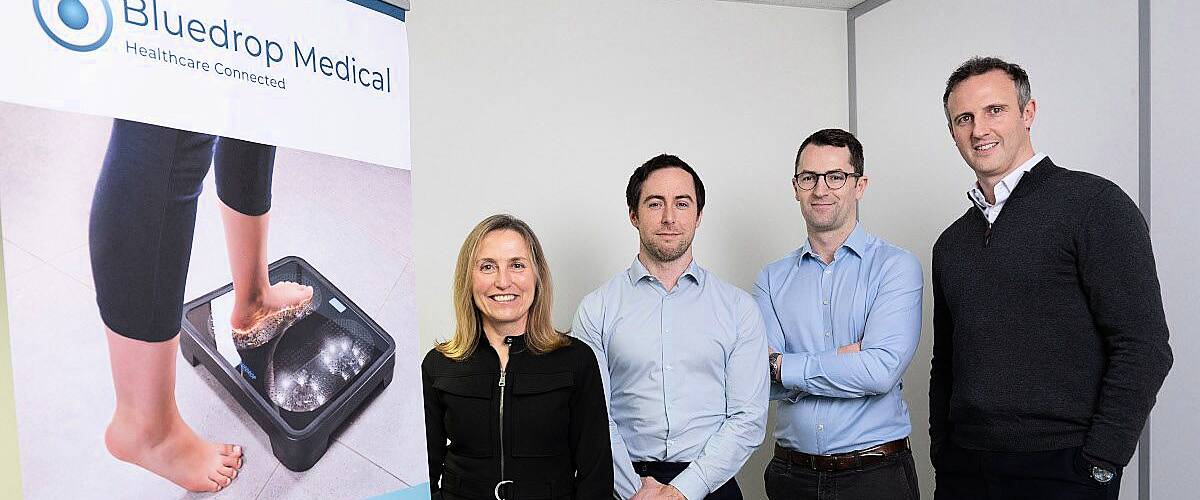 A woman and three men stand in order of their height with a poster on the left that has the Bluedrop Medical logo on it.