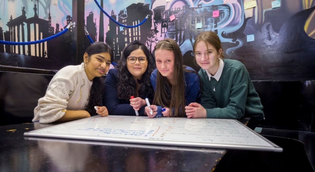 A woman and three teenage girls sitting together at a desk looking at a whiteboard that is laid out in front of them.
