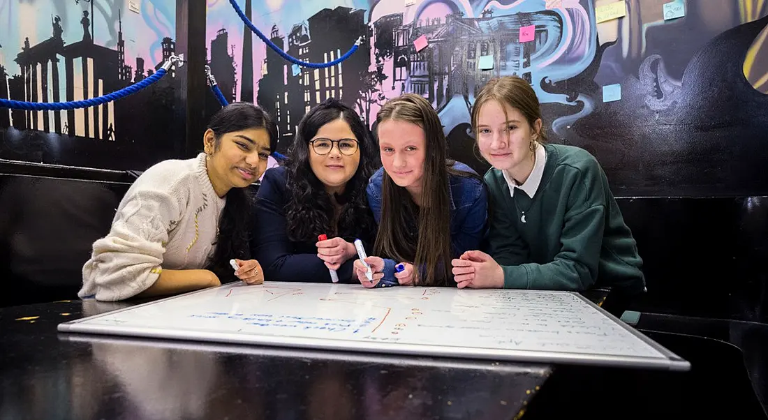 A woman and three teenage girls sitting together at a desk looking at a whiteboard that is laid out in front of them.