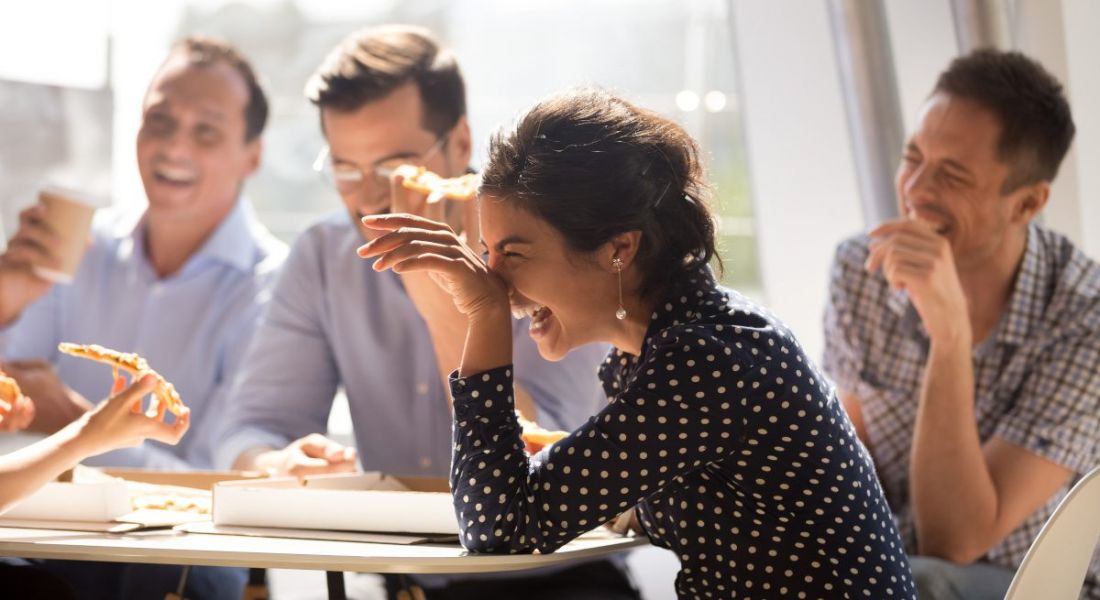Young woman laughing at funny joke eating pizza with diverse coworkers in office.
