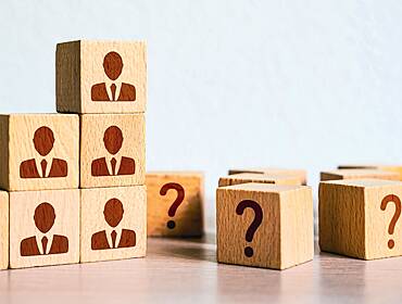 Several wooden blocks stacked on a table with a drawing of a person on each one. Next to this is a scattering of more wooden blocks with question marks.