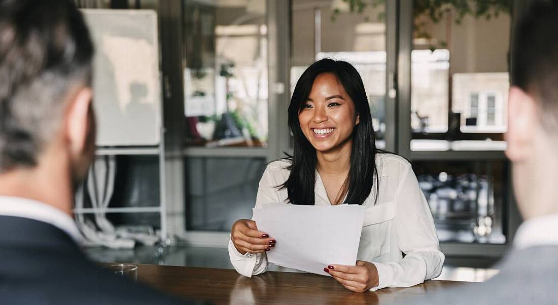 A young woman sitting at a boardroom table across from two men. She is taking part in a job interview.