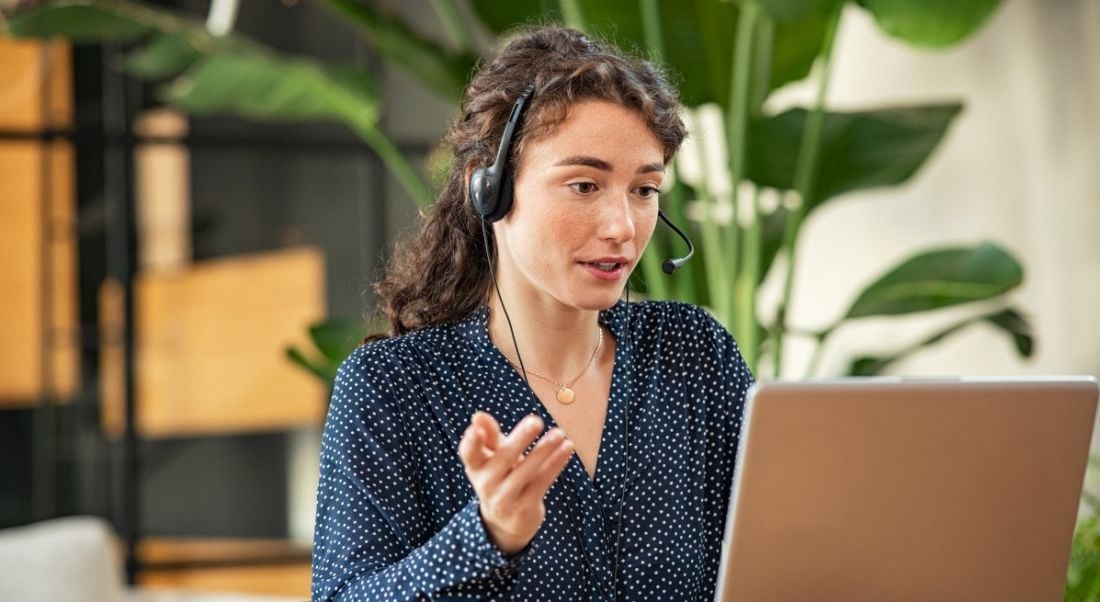 A young woman wearing a headset and talking while working on a laptop, symbolising remote working.