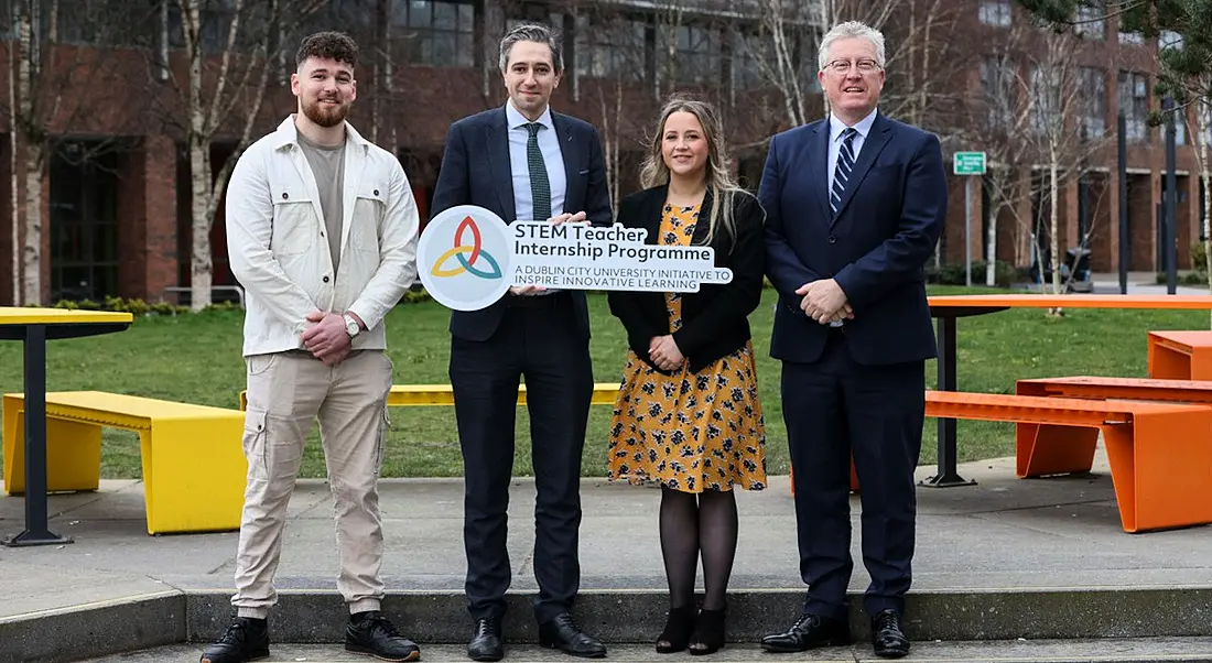 Four people standing outside in front of some benches and a patch of grass holding a sign for teacher internships in STEM.