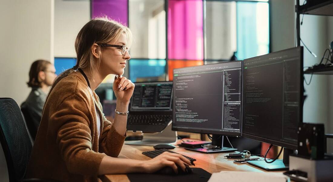 A woman sits at a computer with two screens displaying a lot of code, symbolising women in tech.