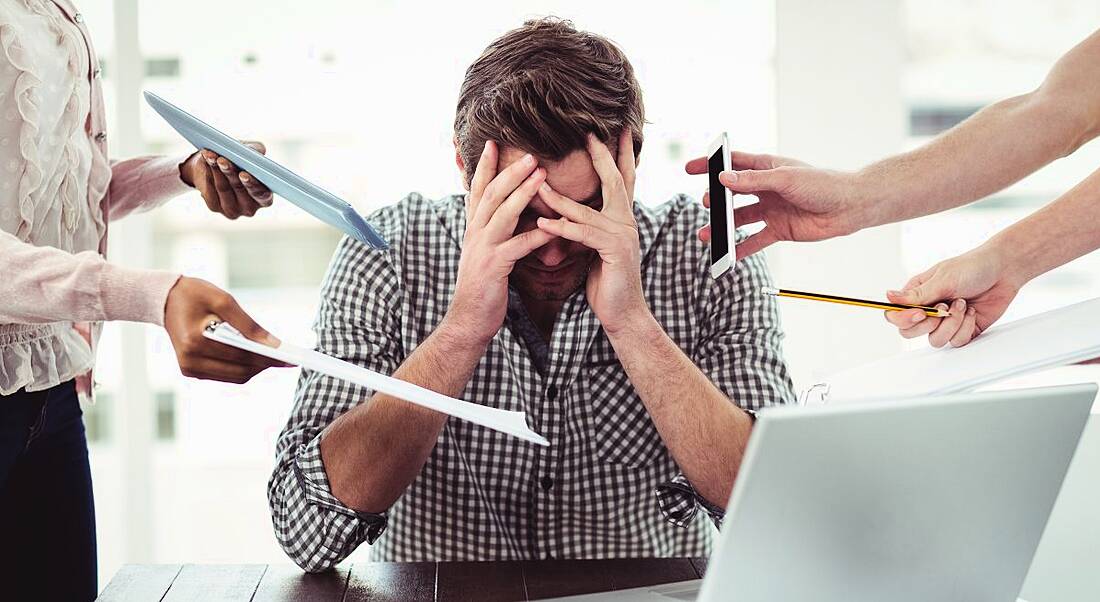 A men in a check shirt sits at a desk with his head in his hands. His laptop is open in front of him and people are standing all around him with papers and phones.