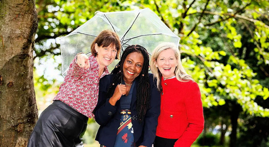 Three women standing under a plastic umbrella under a tree with green foliage. They are smiling at the camera.