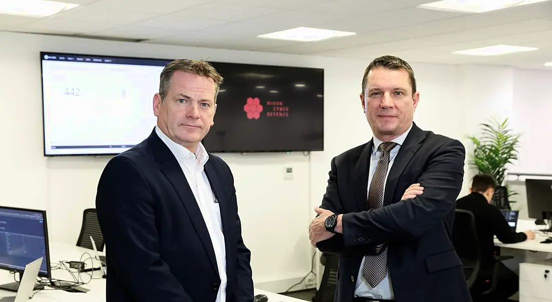 Two men at the Belfast office of Nihon Cyber Defence with company branding on the wall behind them.