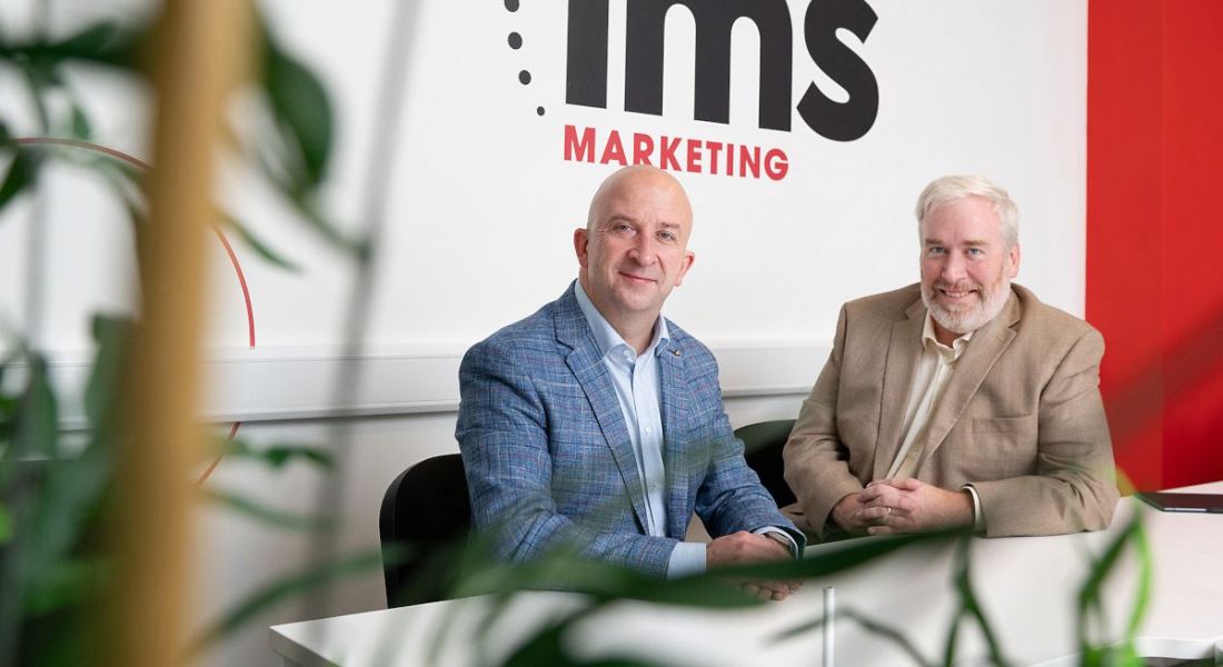 Kevin Moran and Marty Martin of IMS Marketing, a Locomotive Agency sitting in front of a board that has IMS branding on it.